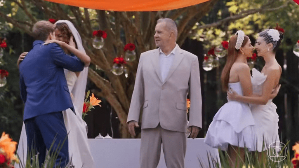 "Em uma cerimônia de casamento ao ar livre, duas noivas vestidas de branco trocam um abraço e um beijo à direita da imagem, enquanto, à esquerda, outra noiva beija um noivo de terno azul. Ao centro, um celebrante observa os casais diante de uma decoração com flores vermelhas e arranjos suspensos. Cena exibida em novela da TV Globo.